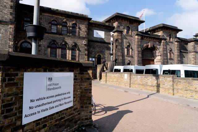 A brick prison building with a sign indicating no vehicle access and several white vans parked nearby.