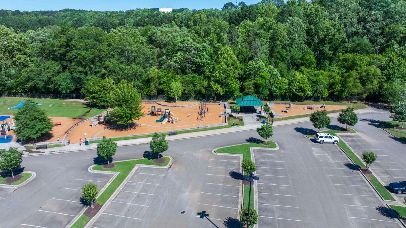 A playground with equipment, seating areas, and a parking lot surrounded by trees.