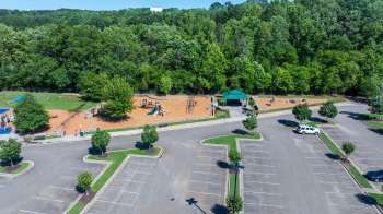 A playground with equipment, seating areas, and a parking lot surrounded by trees.