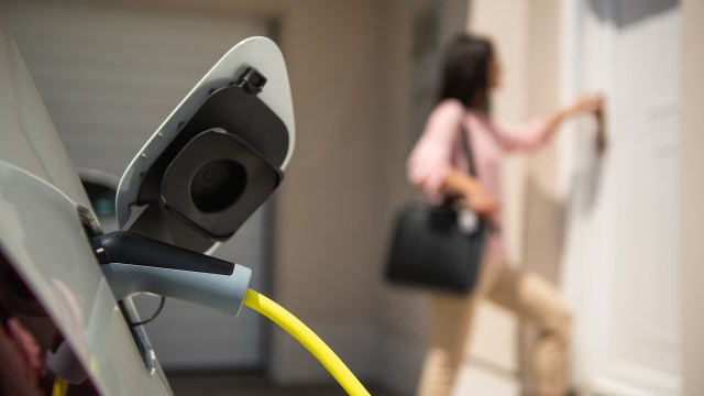 An electric vehicle charging cable is plugged in, while a person approaches a door.