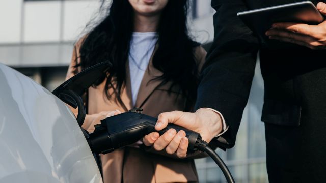 A salesperson connects an electric vehicle charging plug to a car, with another person nearby holding a tablet.