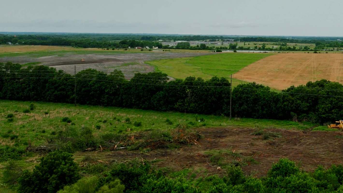 Cleared land with tree stumps and machinery, surrounded by fields and distant trees under a cloudy sky.