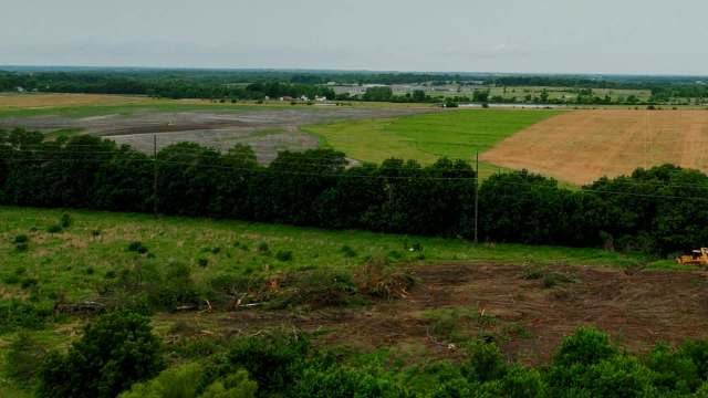 Cleared land with tree stumps and machinery, surrounded by fields and distant trees under a cloudy sky.