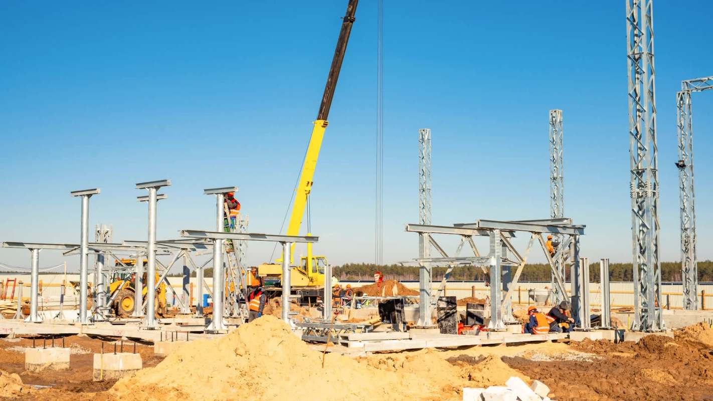 A construction site with various workers assembling metal structures and using machinery under a clear blue sky.