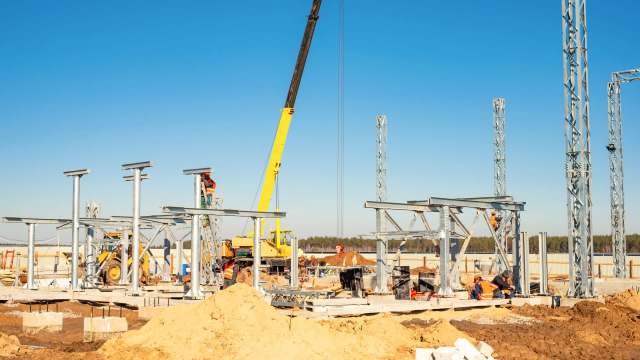 A construction site with various workers assembling metal structures and using machinery under a clear blue sky.