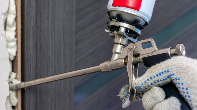 A hand holding a spray gun applying foam sealant to a gap between surfaces.