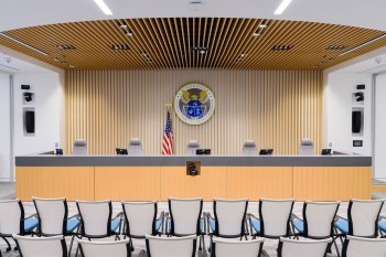 A modern conference room with a FCC seal, American flag, and several empty chairs facing a grey table.