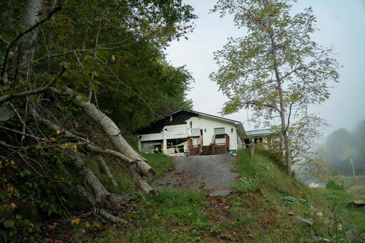 A weathered house sits on a hillside surrounded by trees and overgrown vegetation.