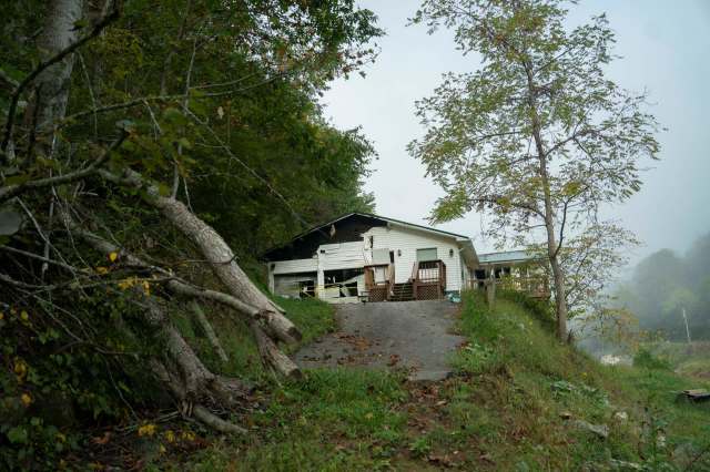 A weathered house sits on a hillside surrounded by trees and overgrown vegetation.