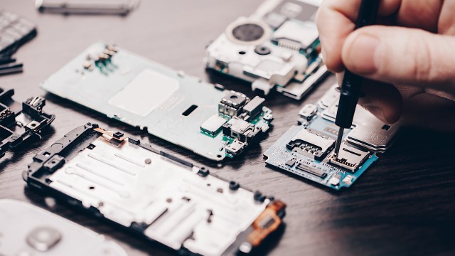 A hand uses a pen tool to work on disassembled smartphone components placed on a table.