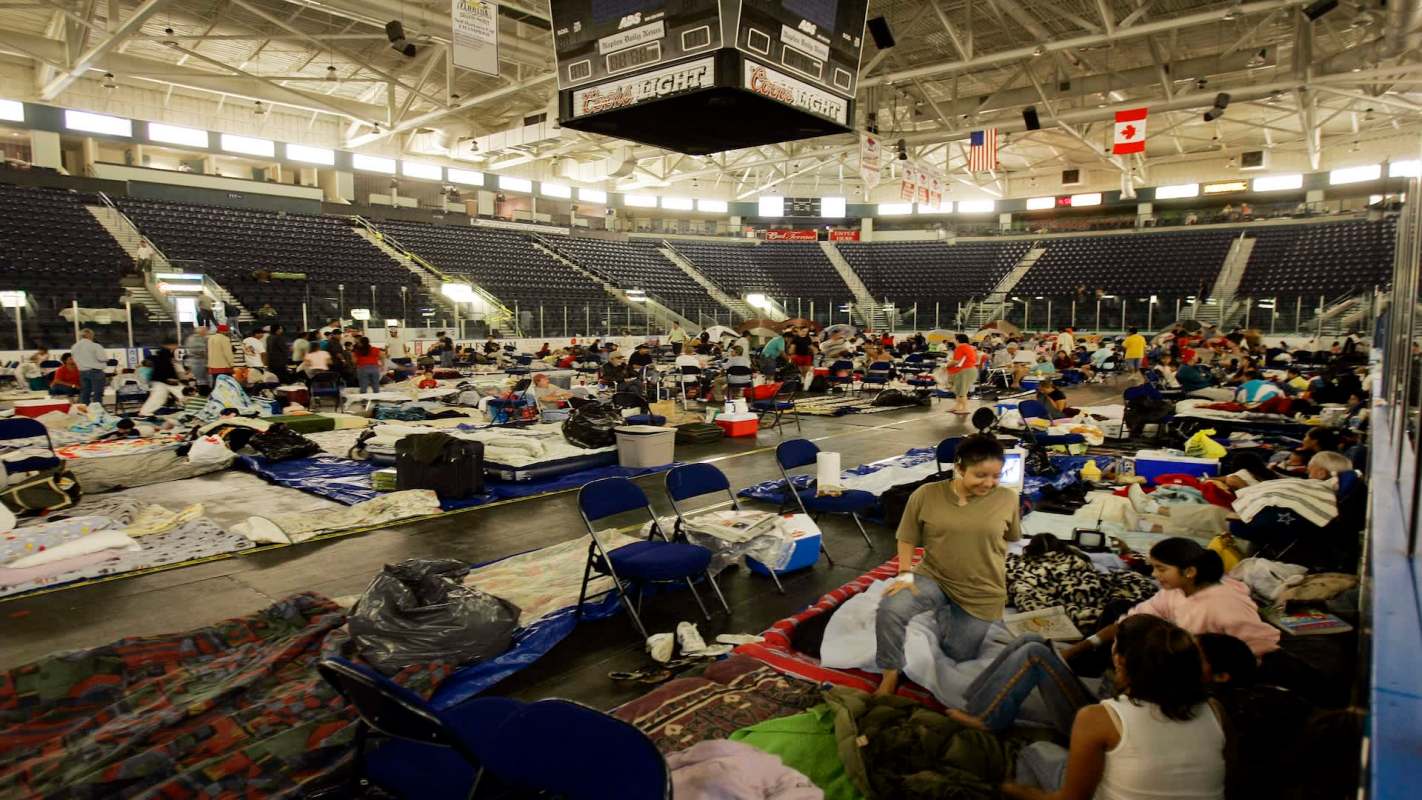 A large indoor space filled with people sitting on blankets and cots, surrounded by personal belongings.