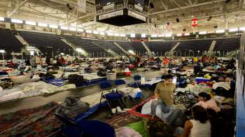 A large indoor space filled with people sitting on blankets and cots, surrounded by personal belongings.