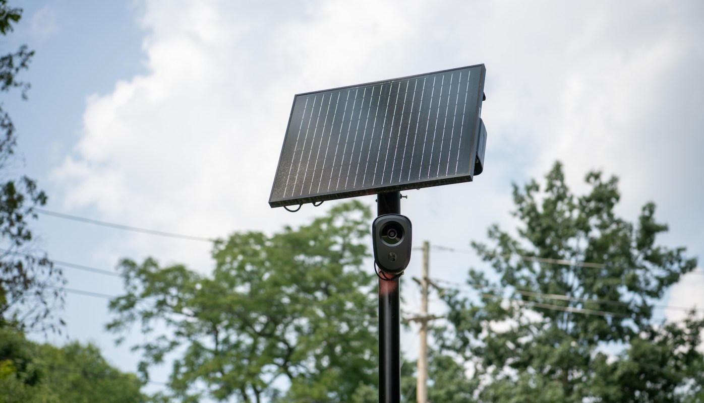 A solar panel mounted on a pole with a camera attached, set against a backdrop of trees and cloudy sky.