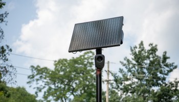 A solar panel mounted on a pole with a camera attached, set against a backdrop of trees and cloudy sky.