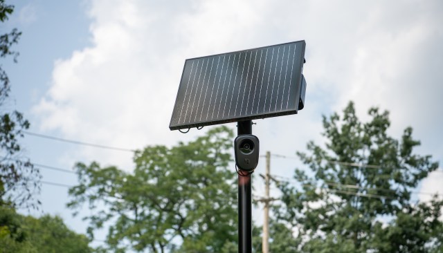 A solar panel mounted on a pole with a camera attached, set against a backdrop of trees and cloudy sky.