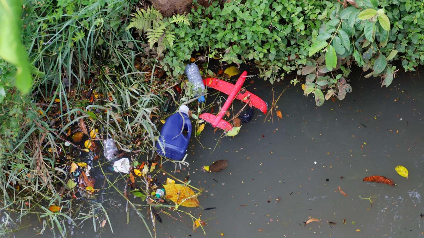 Trash and debris, including plastic bottles and a red toy airplane, accumulate in a shallow stream surrounded by grass and leaves.