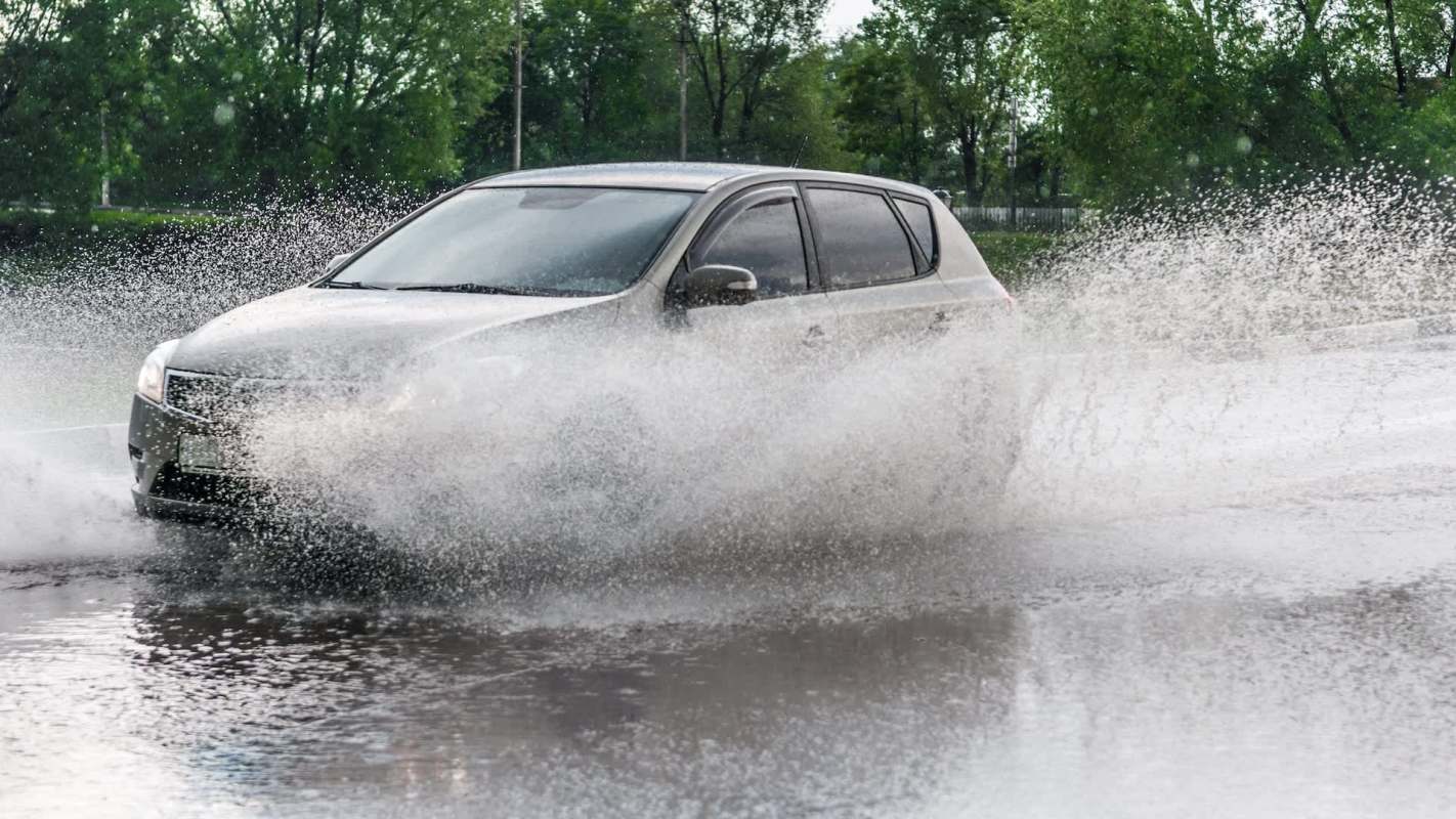 A car drives through a large puddle, spraying water on a wet road.