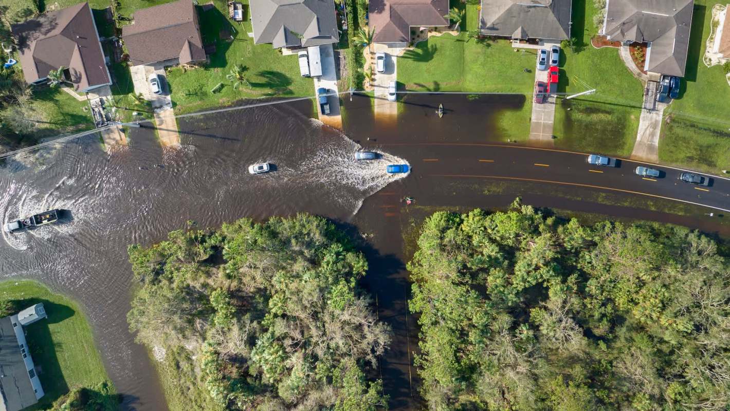 Aerial view of flooded streets with vehicles navigating through water near residential homes and greenery.