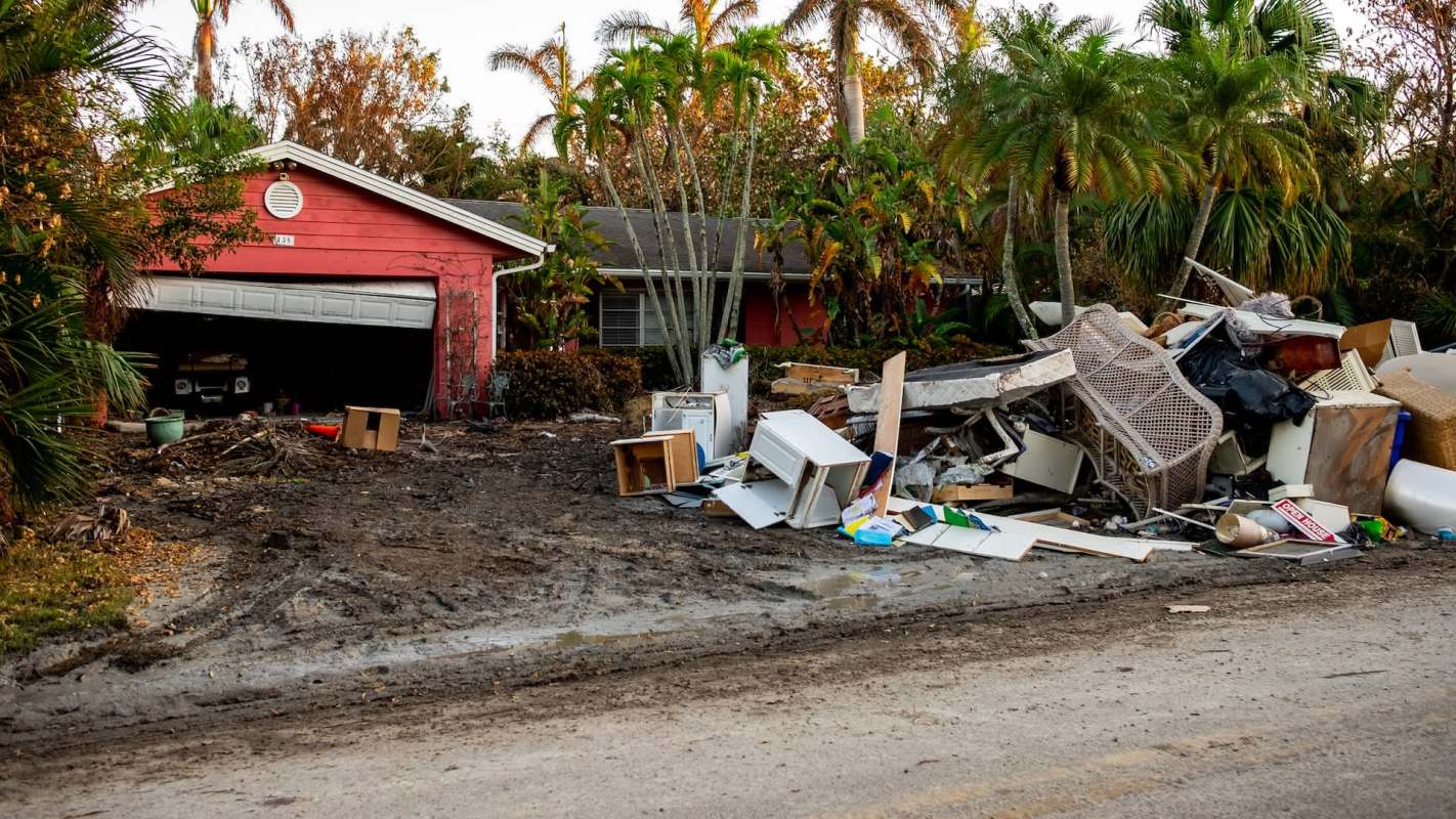 A home with a red exterior and a pile of debris in front, surrounded by palm trees.
