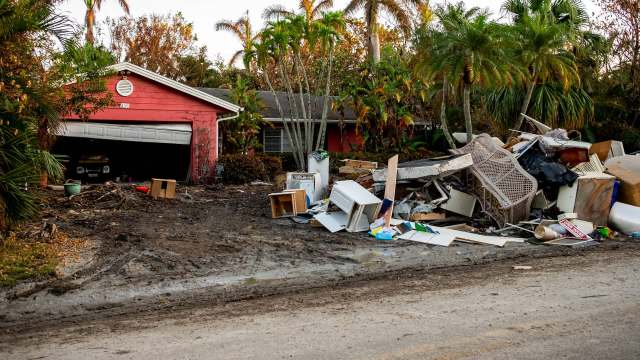 A home with a red exterior and a pile of debris in front, surrounded by palm trees.