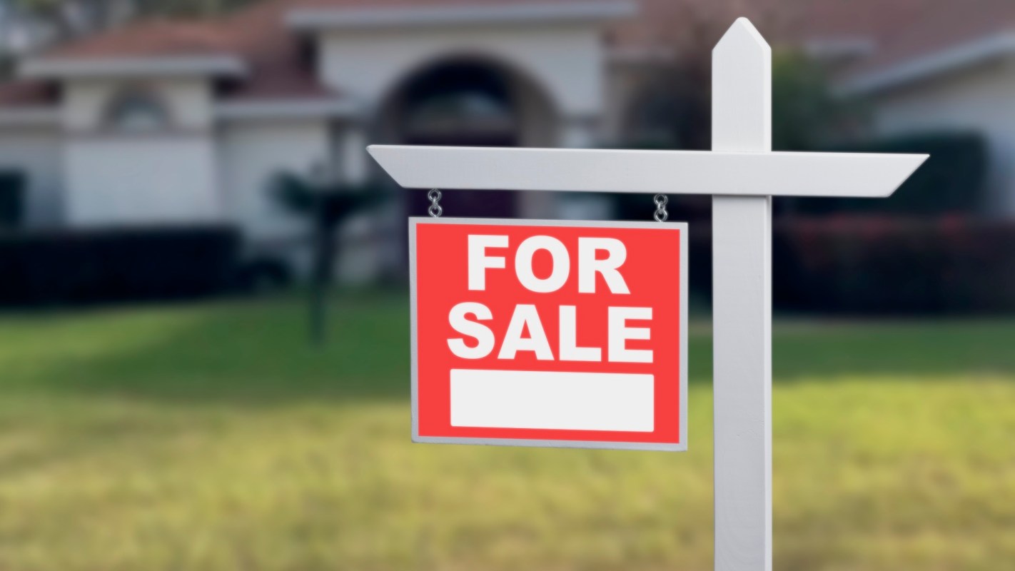 A "For Sale" sign in front of a house with a grassy lawn.