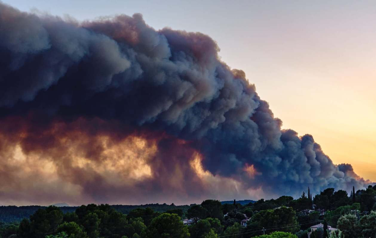 A large plume of smoke rises above a landscape at sunset, obscuring the sky and reflecting orange hues.