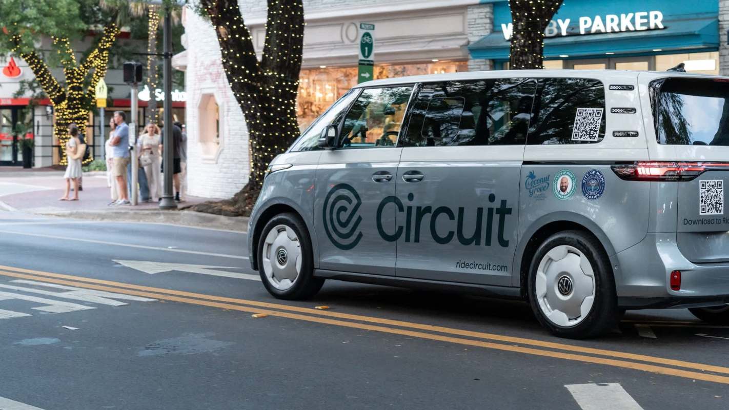 A gray vehicle marked with "Circuit" drives past a group of people on a city street adorned with lights.