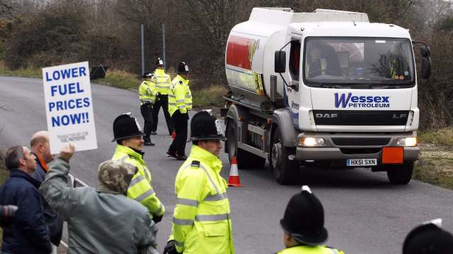 A fuel protest with attendees holding a sign, and police officers monitoring a fuel delivery truck.