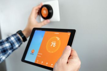A person adjusts a smart thermostat while holding a tablet displaying temperature settings.