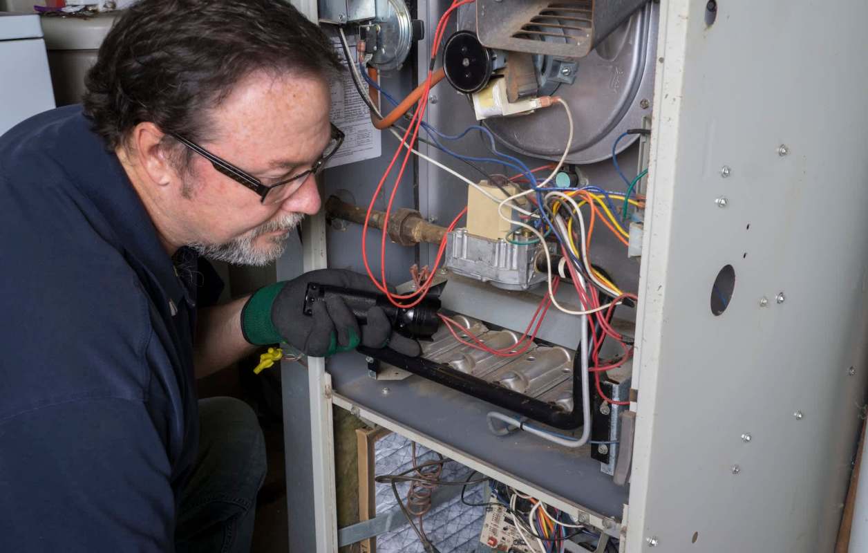 A technician examines the internal components of a furnace with a flashlight.