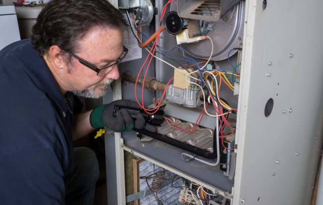 A technician examines the internal components of a furnace with a flashlight.