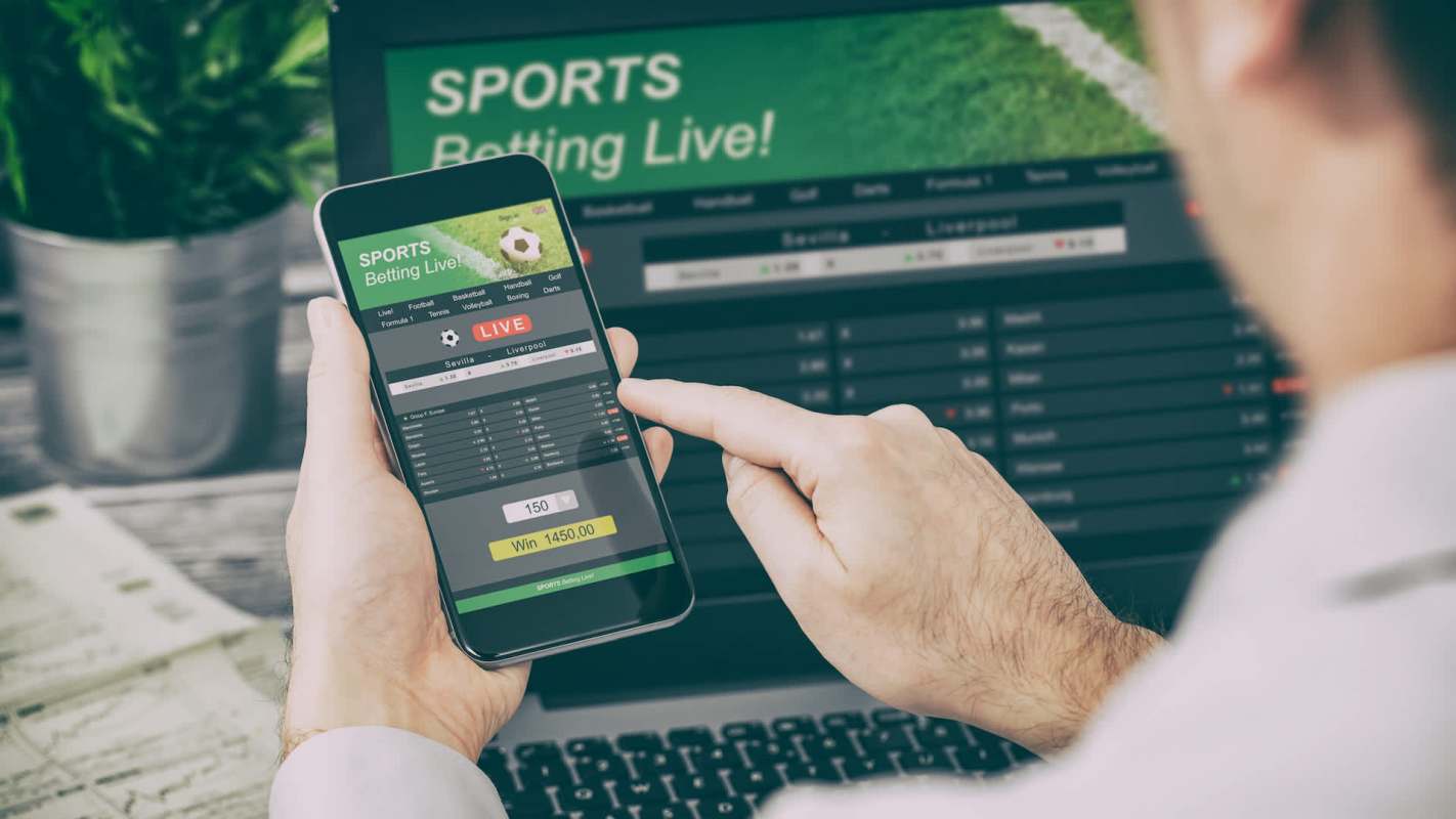 A person interacts with a sports betting app on a smartphone while a laptop displays betting options.