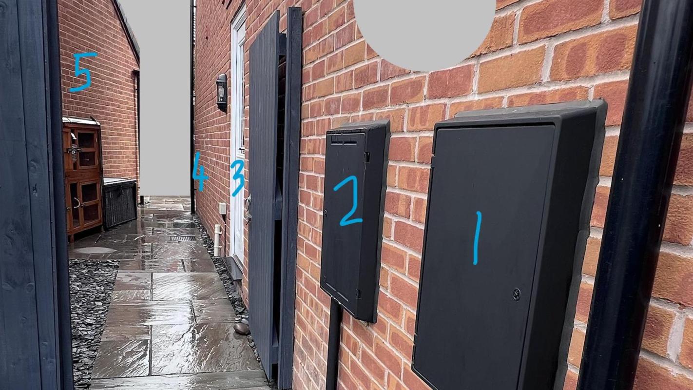 A narrow pathway between brick walls with three black boxes mounted on one side and a wooden cabinet on the other.