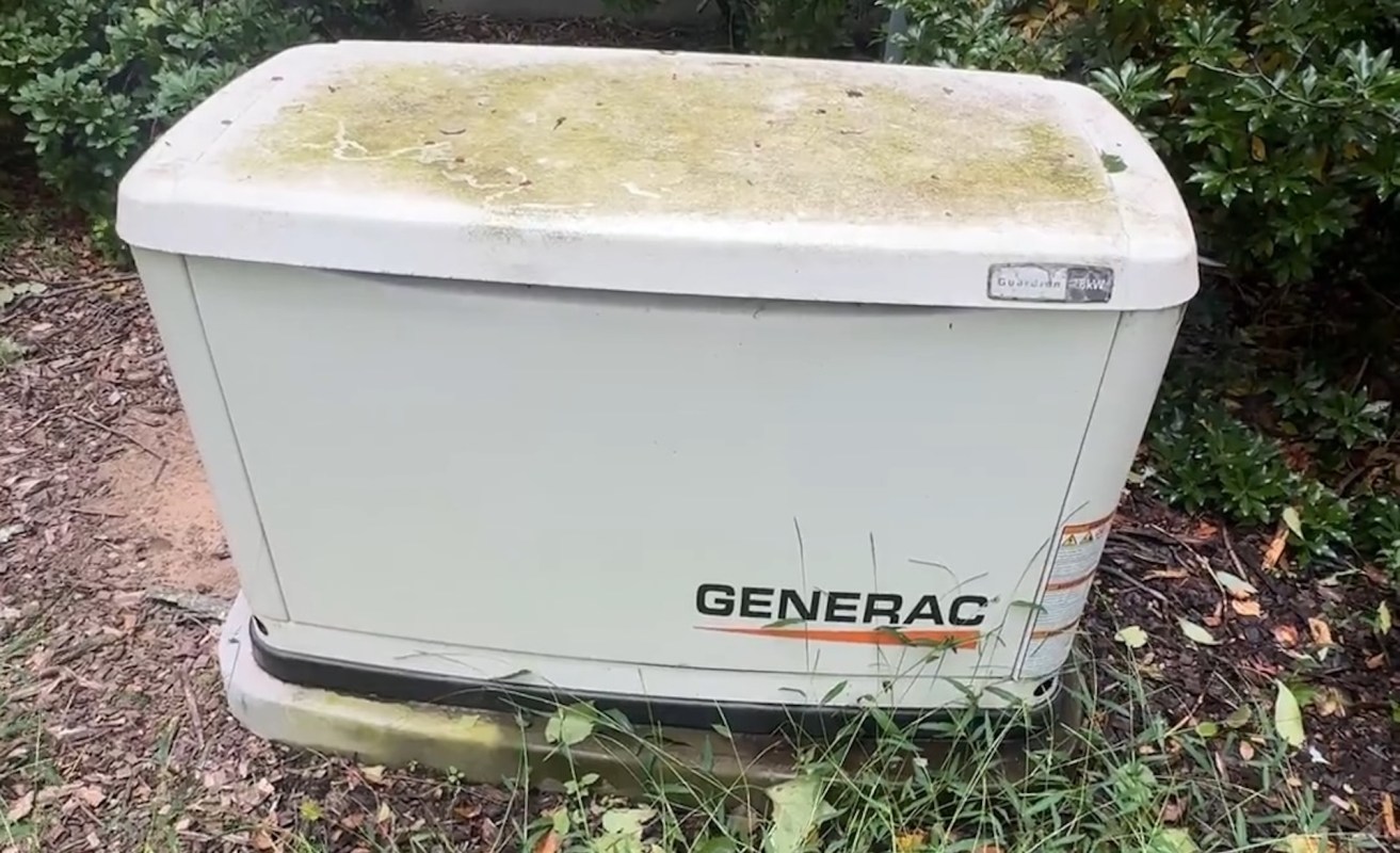 A Generac generator surrounded by overgrown grass and plants.