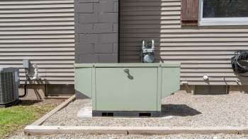 A green generator positioned against a house's exterior wall with gravel landscaping.