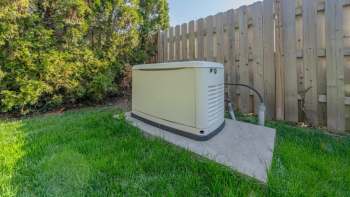 A generator installed on a concrete pad in a grassy area near a wooden fence and bushes.