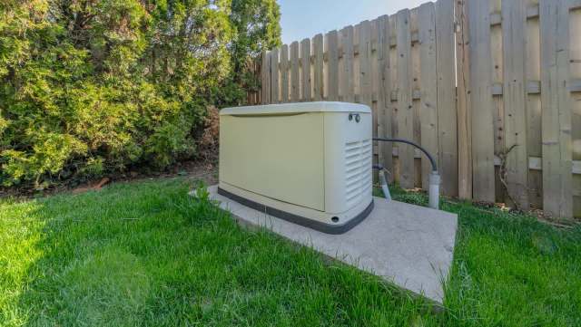 A generator installed on a concrete pad in a grassy area near a wooden fence and bushes.
