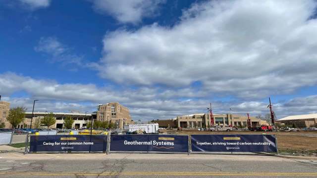 Construction site with a "Geothermal Systems" banner and machinery, under a partly cloudy sky.