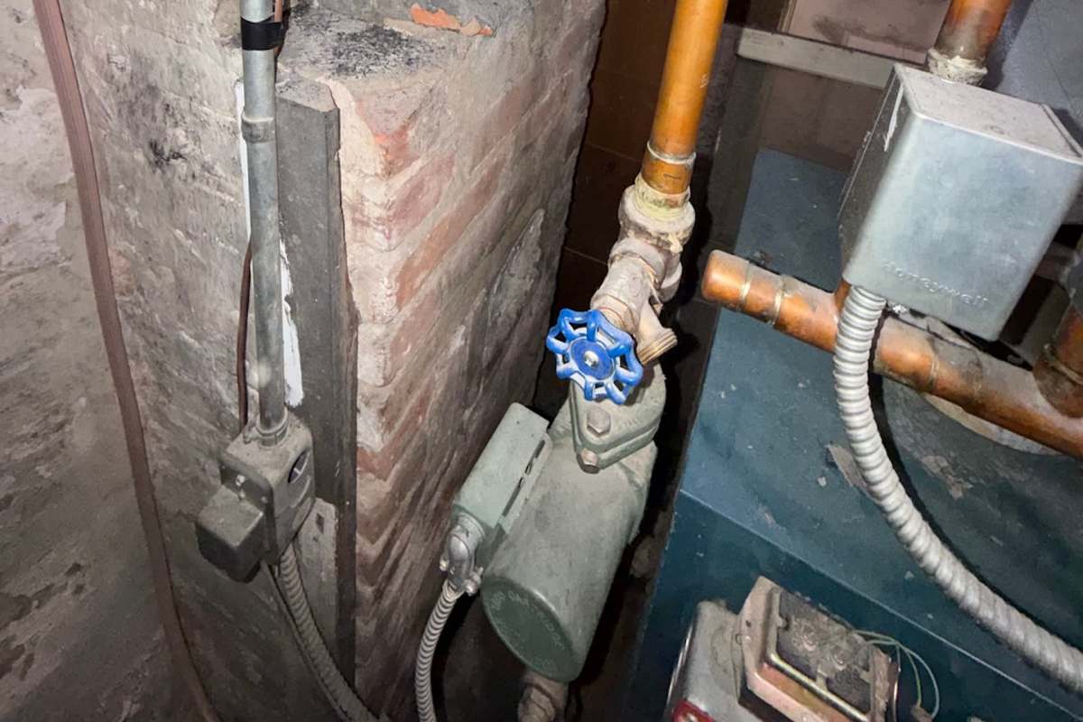 A blue valve and piping system is shown against a brick wall in a basement setting.