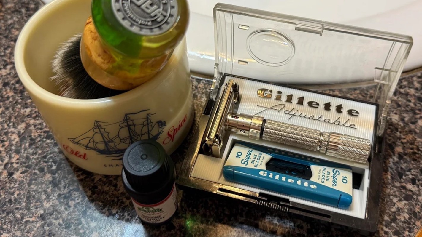 A shaving brush in a mug next to a Gillette adjustable razor and a container of shaving blades.