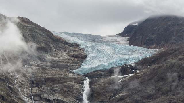 A glacier flows down a mountain, surrounded by rocky terrain and clouds.
