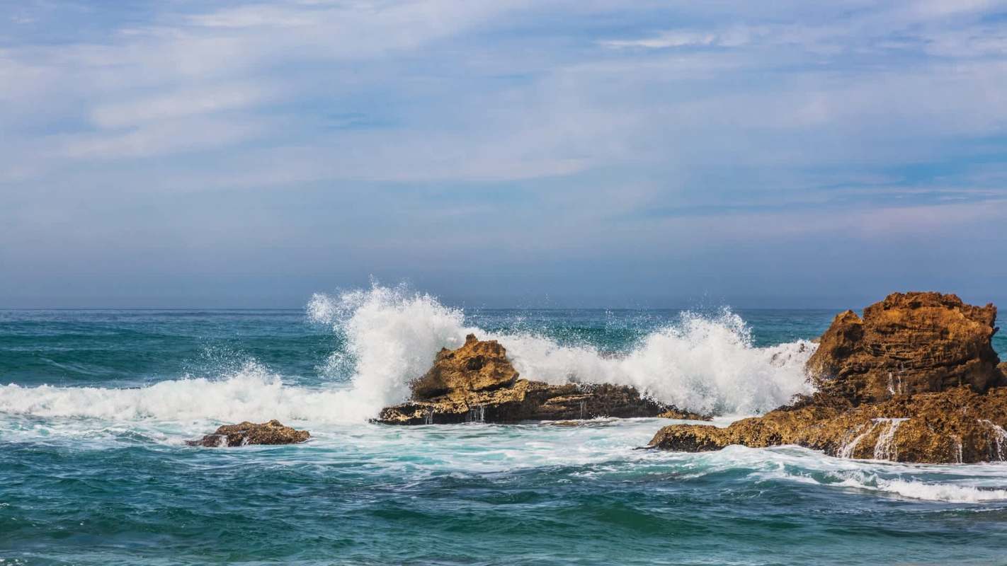 Waves crash against rocky formations in a blue ocean under a partly cloudy sky.