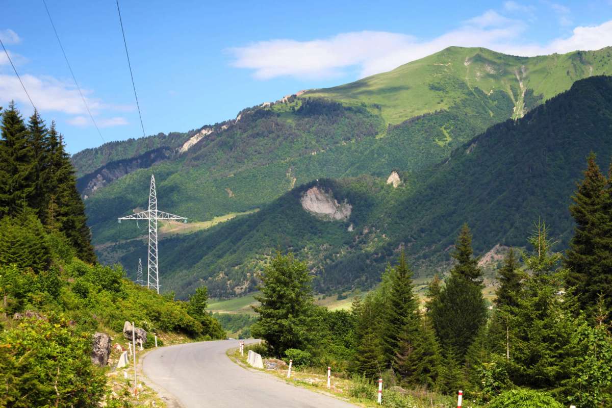 A winding road bordered by greenery and mountains, with a power line in the background.