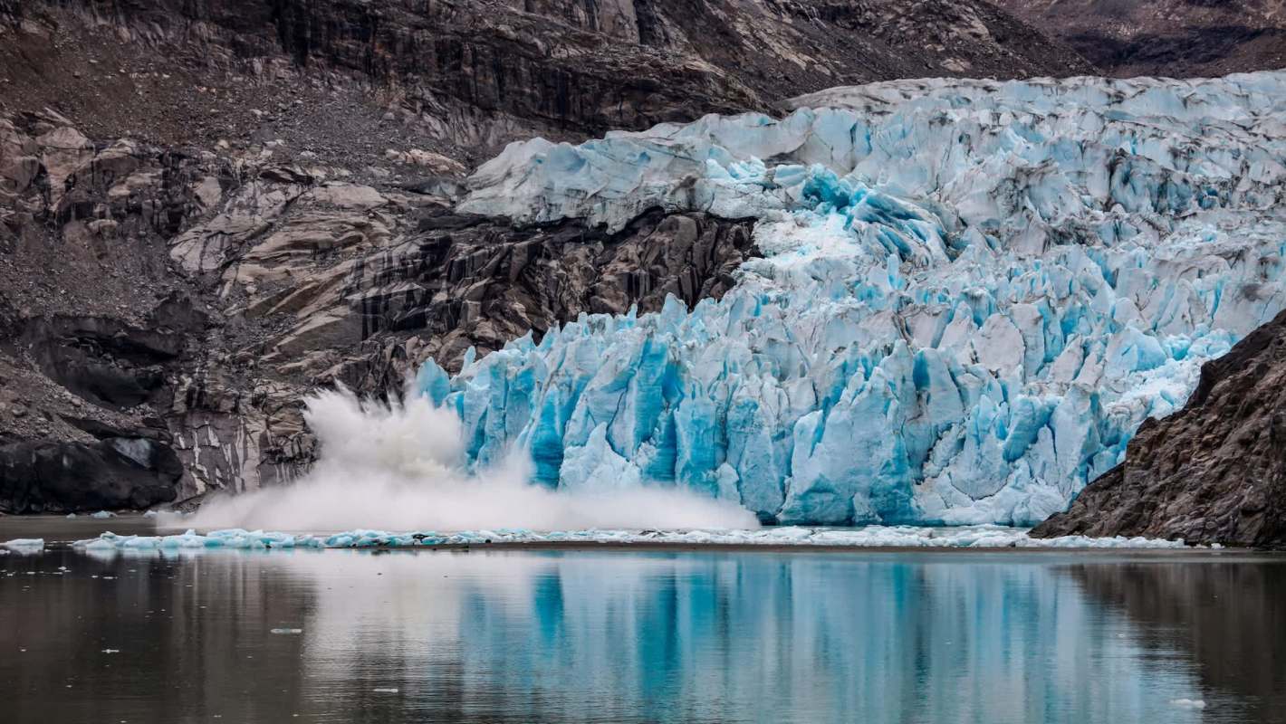 A glacier calving into a calm body of water surrounded by rocky cliffs.