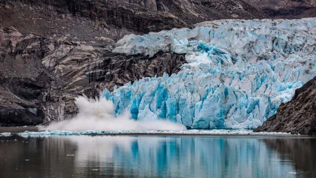 A glacier calving into a calm body of water surrounded by rocky cliffs.