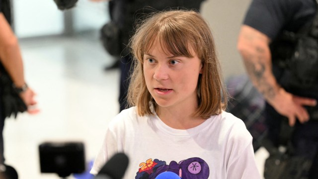 Greta Thunberg speaking into a microphone in a busy public space with people in the background.