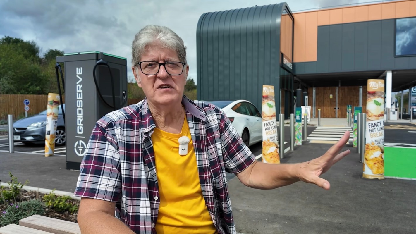 A person gestures towards a building with electric vehicle charging stations and promotional signage.