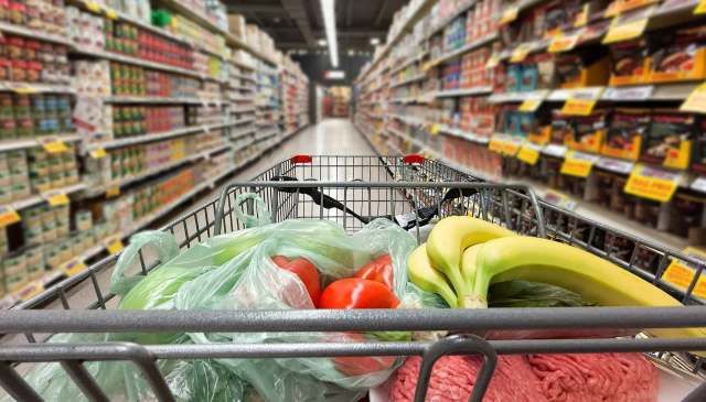 A grocery cart filled with vegetables, fruit, and packaged goods in a supermarket aisle.