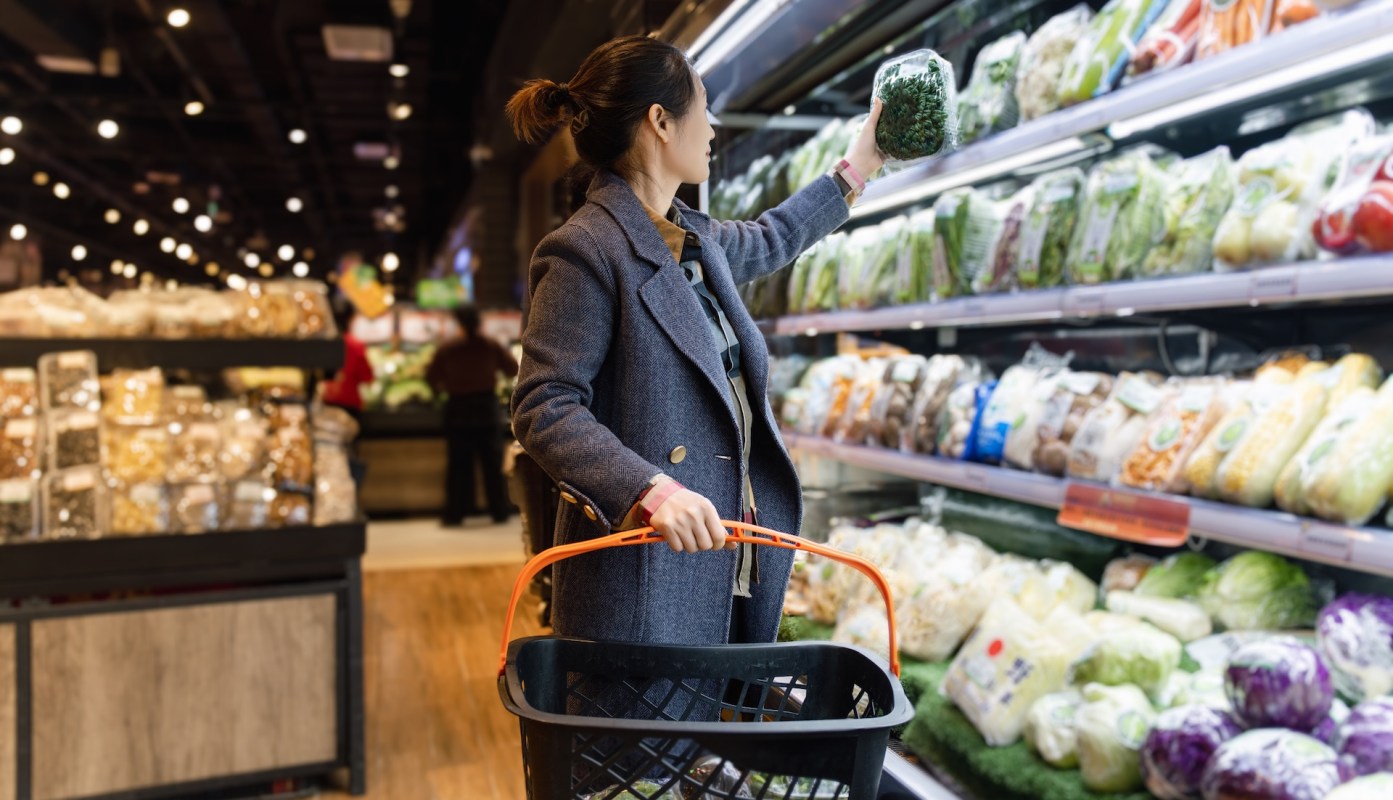 A woman reaches for a jar of greens in a grocery store aisle filled with various vegetables.