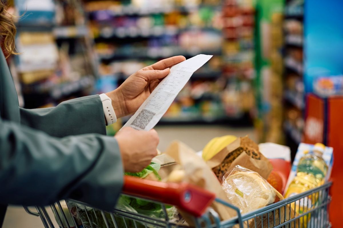 A person examines a receipt while standing next to a grocery cart filled with various food items.